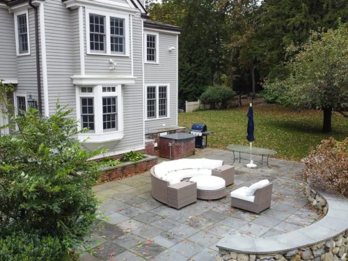 A backyard patio with a circular seating area, grill, and table, surrounded by trees and greenery.