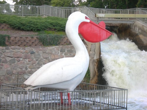 A large white sculpture of a pelican with a red beak stands near a waterfall, surrounded by a metal fence.