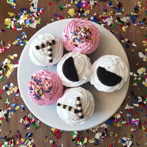 A plate of colorful cupcakes with pink frosting, sprinkles, and chocolate decorations, surrounded by confetti.