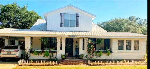 A charming white house with a metal roof, front porch, and lush greenery, featuring a car parked nearby.