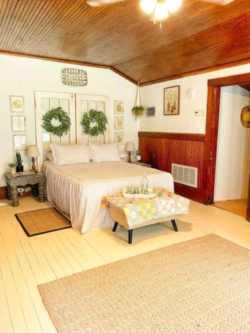 Cozy bedroom with a bed, decorative wreaths, wooden accents, and a colorful quilt on a small table.