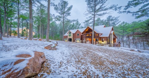A cozy wooden cabin surrounded by snow-covered trees and a gravel driveway in a winter landscape.