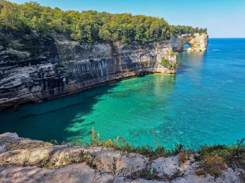 Scenic view of a rocky coastline with clear turquoise water and lush greenery under a bright blue sky.
