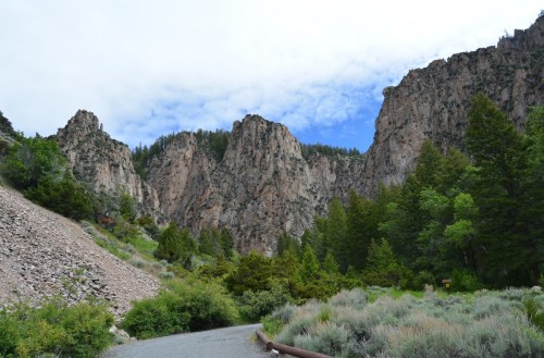 A winding road leads through a rocky canyon with tall cliffs and lush green trees under a partly cloudy sky.