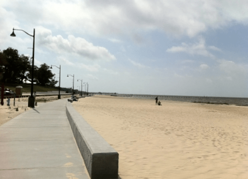 A sandy beach with a walkway, lined by lampposts, under a partly cloudy sky. People are seen in the distance.