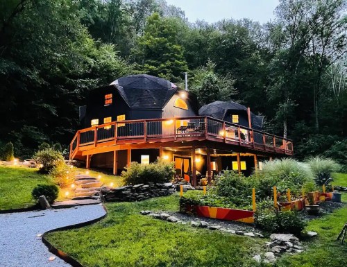 A modern black dome house with a wooden deck, surrounded by lush greenery and illuminated by soft outdoor lights.