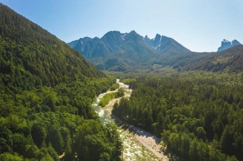 Aerial view of a winding river surrounded by lush green forests and majestic mountains under a clear blue sky.