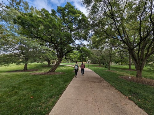 Two people walk along a paved path surrounded by lush green trees and grass under a partly cloudy sky.