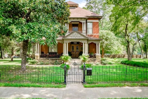 A large, historic brick house with a porch, surrounded by greenery and a black iron fence.