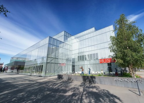 Modern glass building with angular design, featuring the word "MUSE" and surrounded by trees in Anchorage.
