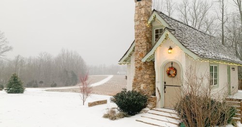 A cozy cottage with a stone chimney, surrounded by snow and trees, decorated with lights and a wreath on the door.