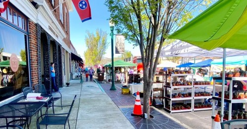 A vibrant street market scene with colorful tents, shoppers, and Tennessee flags, lined with trees and outdoor seating.