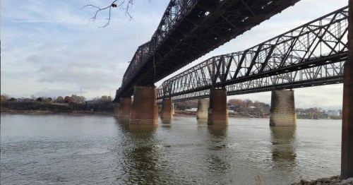 A view of two large railway bridges spanning a river, with reflections on the water and a cloudy sky above.