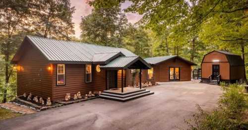 Two brown cabins with green roofs surrounded by trees, featuring a paved driveway and landscaped garden.