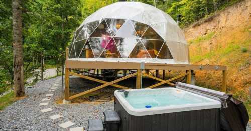 A geodesic dome cabin on a wooden deck, surrounded by trees, with a hot tub in the foreground and a gravel path leading up.