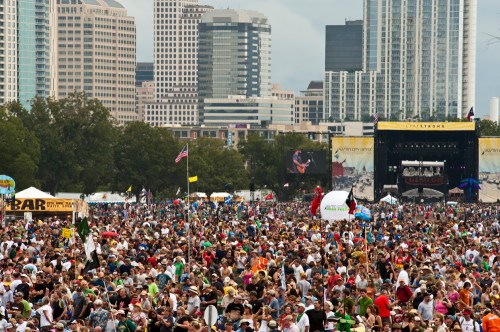 A large crowd gathers at an outdoor festival with city skyscrapers in the background and a stage visible.