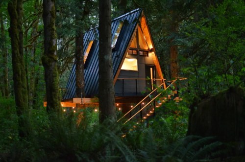 A modern A-frame cabin nestled in a lush forest, illuminated warmly at night with steps leading up to it.