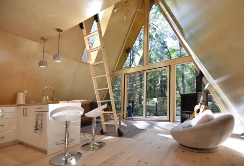 A modern A-frame cabin interior with large windows, a ladder, and minimalist furniture, surrounded by trees.