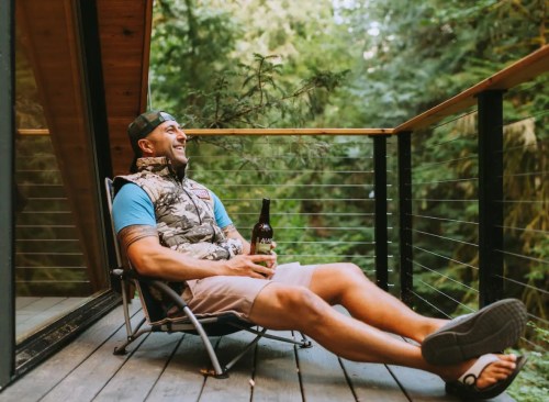 A man relaxes in a chair on a deck, surrounded by lush greenery, holding a bottle and enjoying the outdoors.