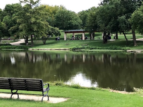 A peaceful park scene with a bench by a pond, surrounded by green trees and a gazebo in the background.