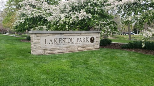 Sign for Lakeside Park, surrounded by green grass and flowering trees, indicating a public recreational area.