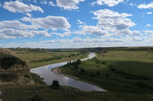 A winding river flows through a green valley under a blue sky with fluffy clouds. Scenic landscape with hills in the background.