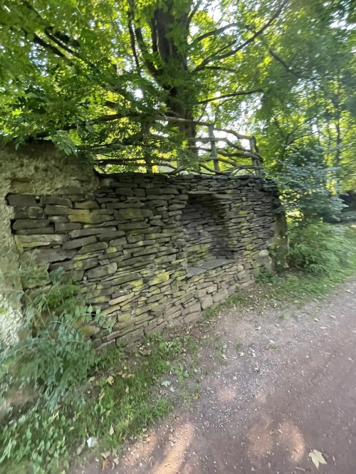 A stone wall partially covered by greenery, with a wooden fence above, along a dirt path in a wooded area.