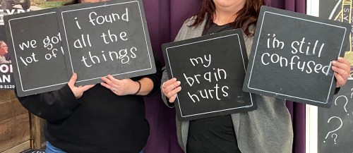 Two women hold chalkboards with playful messages: "we got a lot of," "i found all the things," "my brain hurts," and "i'm still confused."