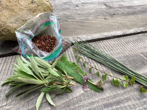 A plastic bag of seeds next to fresh herbs and a rock on a wooden surface.