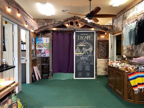 Interior of a cozy shop featuring a rustic design, chalkboard with "Escape Riddle," and various merchandise displays.