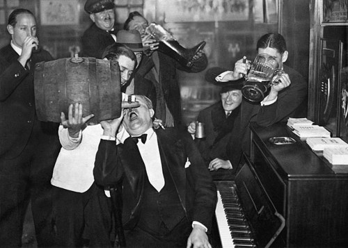 A lively group of men in formal attire enjoying drinks and music around a piano in a vintage setting.