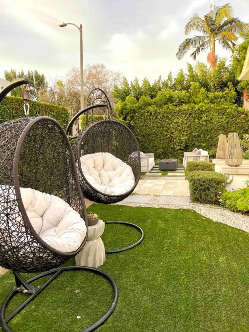 Two hanging chairs with cushions on a green lawn, surrounded by hedges and decorative planters.