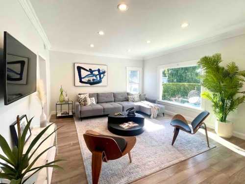 Bright living room with a gray sofa, round coffee table, plants, and large windows letting in natural light.