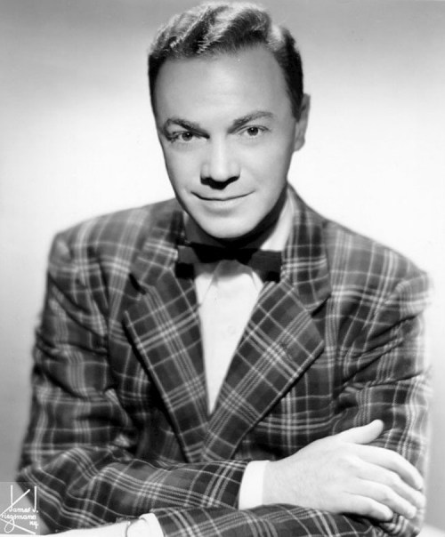 A black-and-white portrait of a man in a plaid suit and bow tie, smiling and posing with crossed arms.