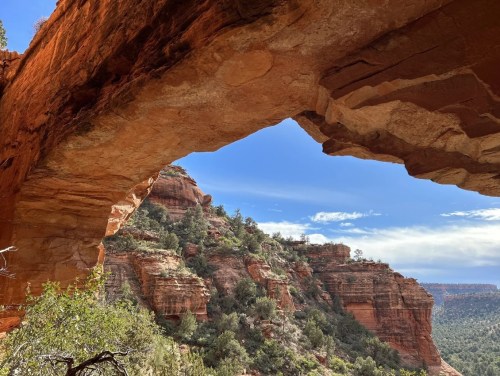 A natural rock arch frames a scenic view of red cliffs and green trees under a blue sky.