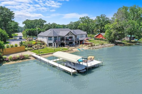 Aerial view of a lakeside home with a dock, surrounded by greenery and a clear blue sky.