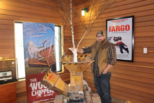 A man with a beard stands next to a wood chipper in a wooden room, with promotional materials and a movie poster in the background.