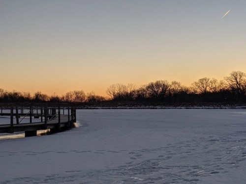 A serene winter scene with a frozen lake, a wooden dock, and trees silhouetted against a colorful sunset sky.