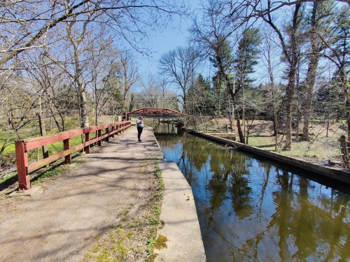 A person walks along a path beside a calm waterway, with trees and a red bridge in the background on a sunny day.