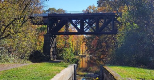 A black metal bridge spans a calm canal, surrounded by vibrant autumn foliage and clear blue skies.
