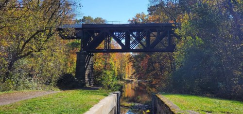 A metal bridge spans a tranquil canal, surrounded by colorful autumn foliage and clear blue skies.
