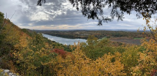 A panoramic view of a river surrounded by autumn foliage and rolling hills under a cloudy sky.
