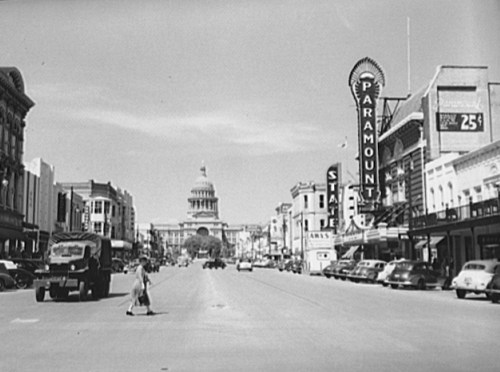 Black and white image of a busy street with vintage cars, a theater marquee, and a government building in the background.