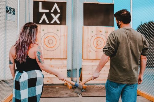 A man and woman hold axes, facing two wooden targets in an axe-throwing venue. The backdrop features "LAX."