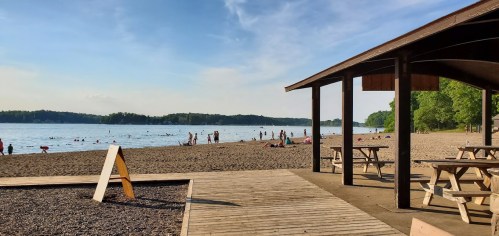 A sandy beach with people swimming and relaxing, framed by trees and a wooden pavilion under a clear blue sky.