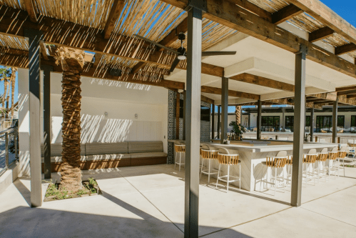 A modern outdoor space with a shaded seating area, bar stools, and palm trees under a clear blue sky.