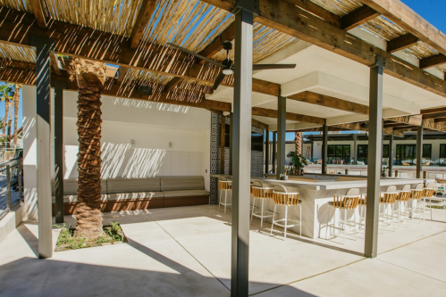 A modern outdoor space with a shaded seating area, bar stools, and palm trees under a clear blue sky.