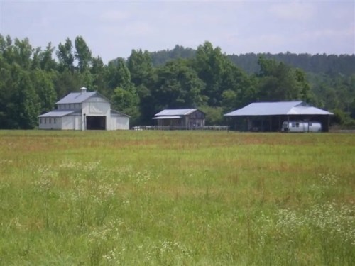 A rural scene featuring two barns and a field, surrounded by trees under a clear blue sky.