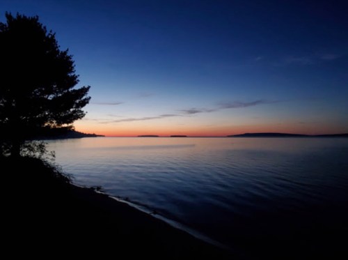 A serene lakeside view at dusk, with a silhouette of trees and a gradient sky transitioning from blue to orange.