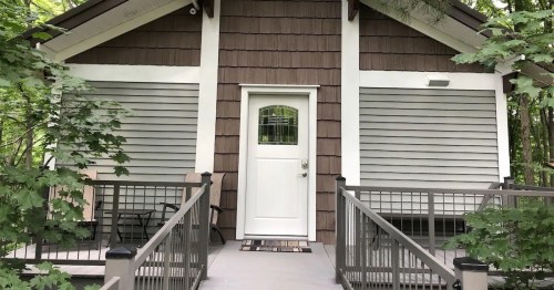 A cozy cabin with brown and gray siding, featuring a white door and surrounded by greenery.